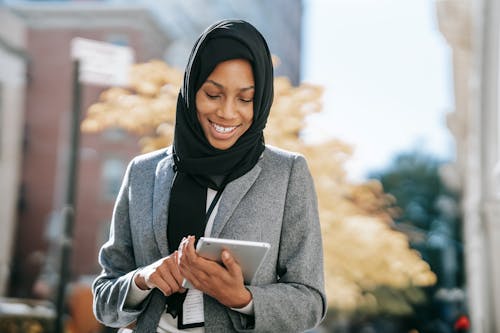 Positive African American female in gray coat and hijab with badge messaging on tablet