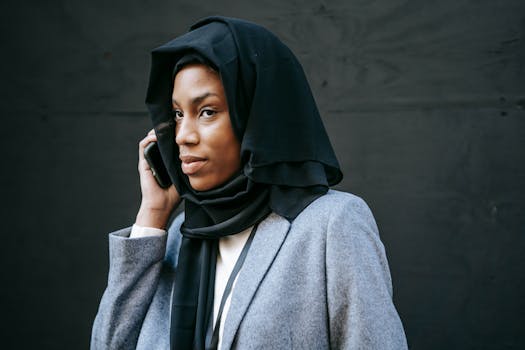 Serious young African American female in hijab and gray coat speaking on phone and looking away against black background