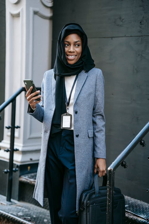 Positive young African American female in gray coat and hijab with bag and smartphone looking away while standing on stairs