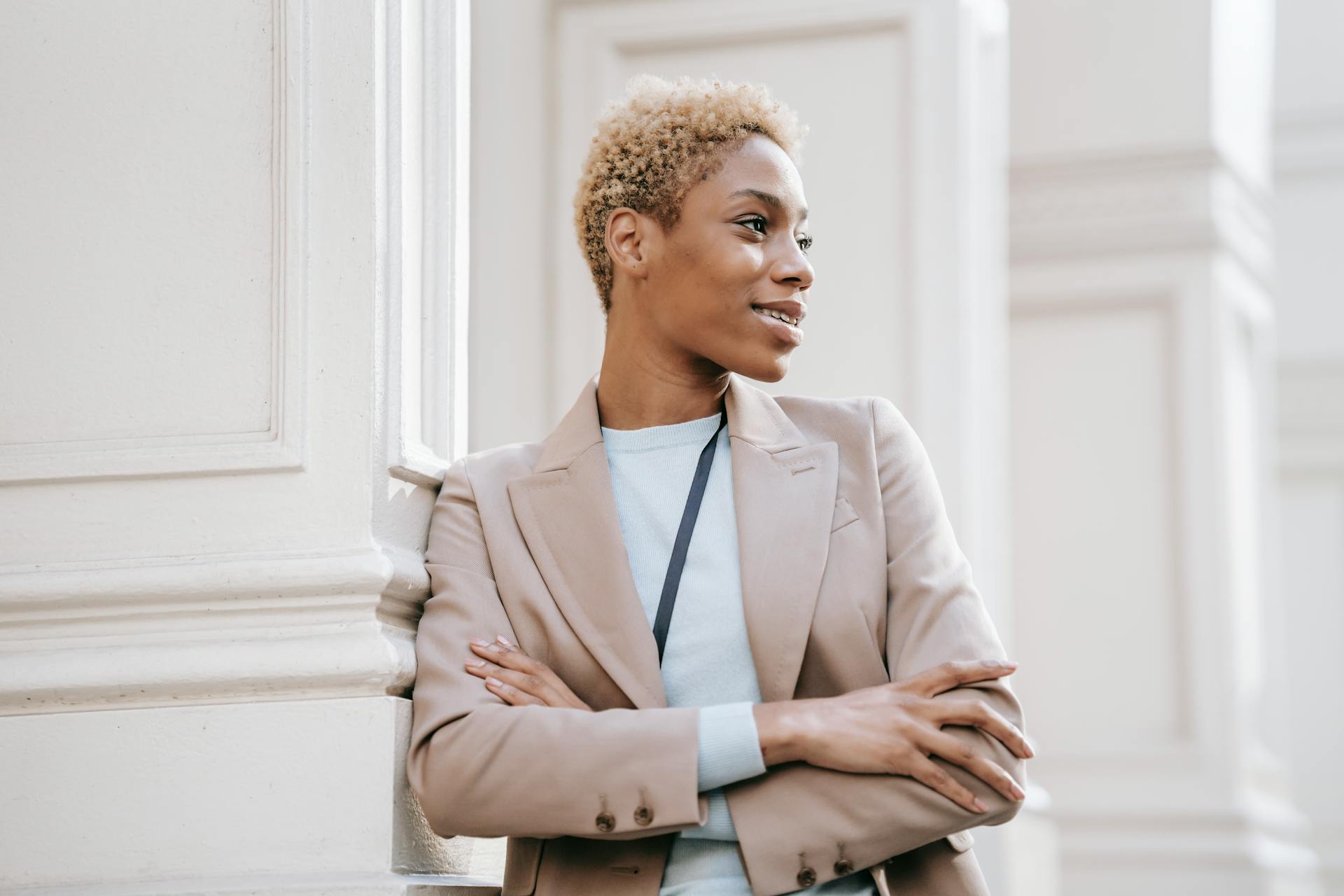 Young dreamy ethnic female executive in formal wear with crossed arms looking away near ornamental column in city