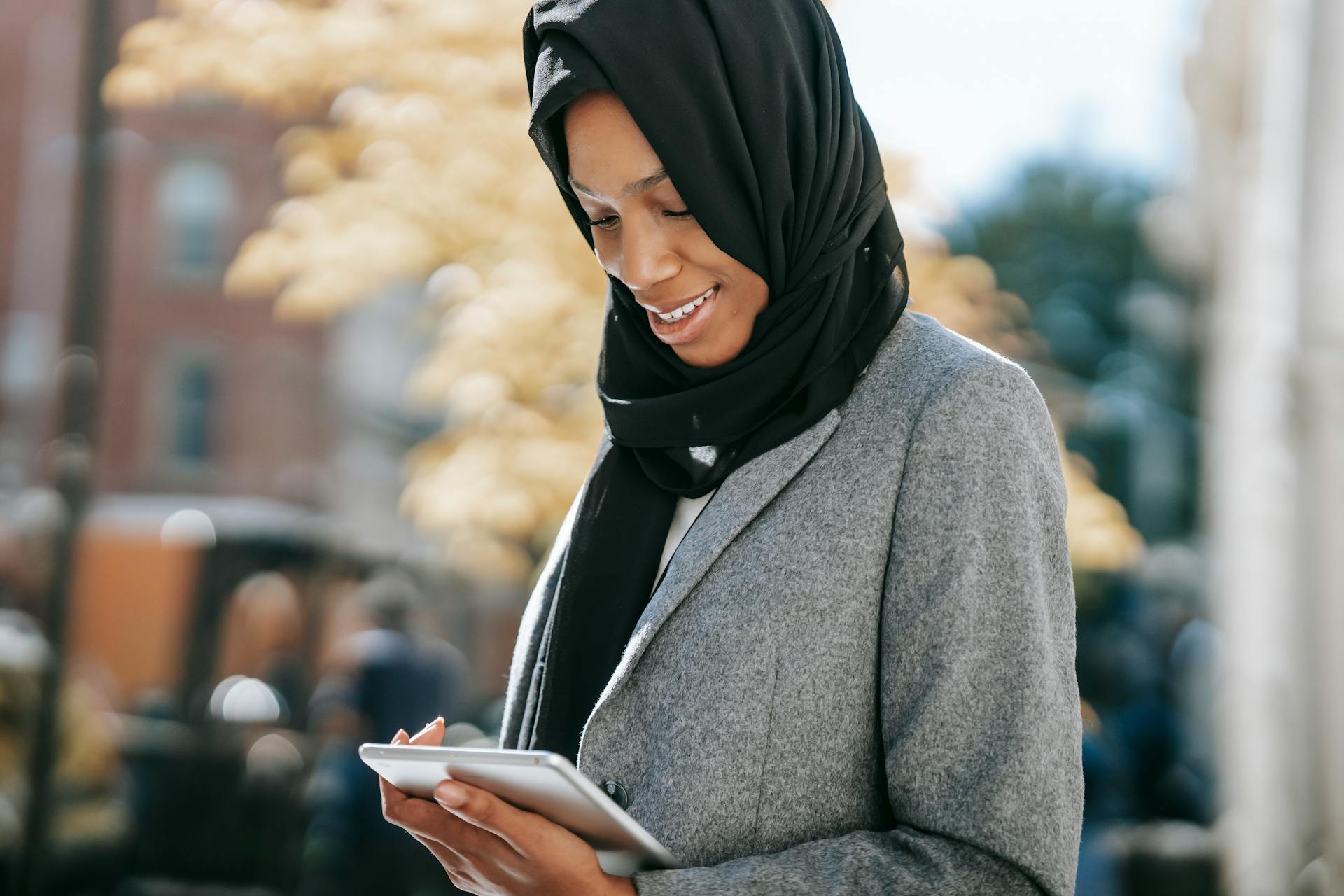 Content young African American Muslim businesswoman in stylish suit and traditional hijab smiling and reading notes on tablet while standing on city street