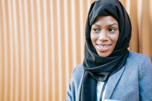Free Young positive ethnic Muslim lady in elegant suit and traditional hijab smiling and looking away while standing near corrugated metal wall on street Stock Photo