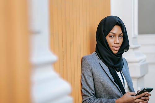 Gorgeous young ethnic Muslim female entrepreneur in classy suit and traditional headscarf standing near building on street and using smartphone