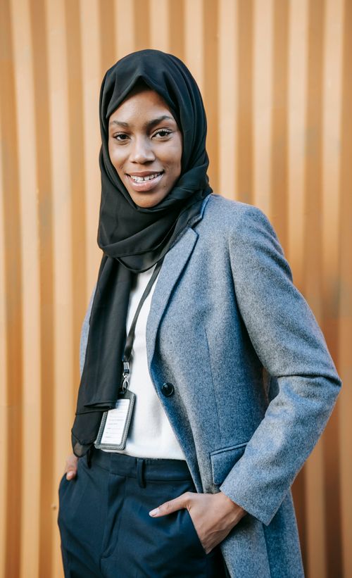 Free Confident young African American Islamic businesswoman in elegant suit and traditional hijab smiling and looking at camera while standing near corrugated metal wall with hands in pockets Stock Photo