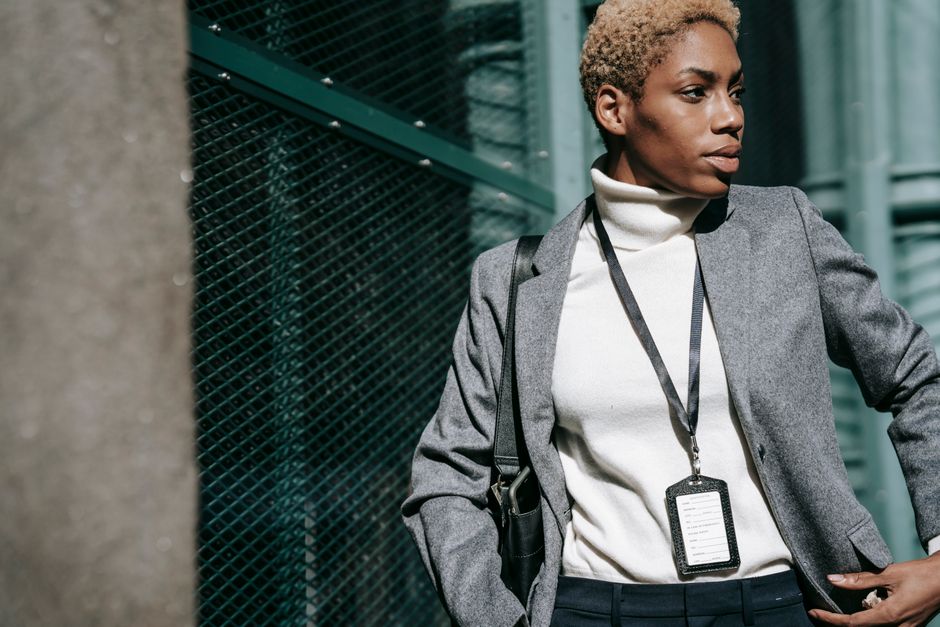 Confident young African American female entrepreneur with short blond hair in elegant outfit with name tag on neck standing on street and looking away thoughtfully on sunny day