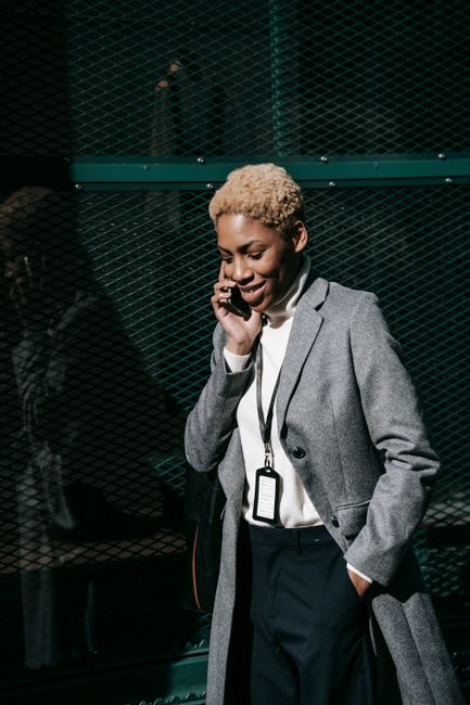 Self assured young black businesswoman with short hair and in stylish elegant outfit walking on street and smiling while having phone conversation on sunny day