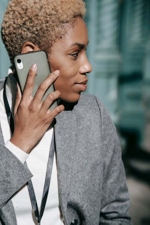 Crop happy young African American lady with short blond hair in classy outfit smiling while talking on mobile phone on street