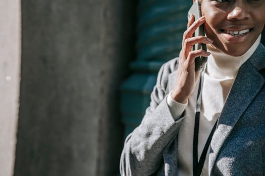 Crop cheerful young African American woman in elegant clothes smiling while talking on smartphone on street on sunny day