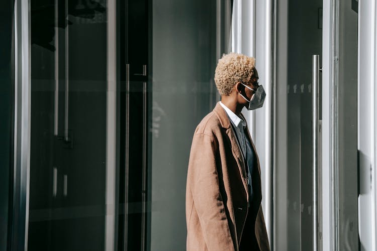 Black Woman In Mask Standing Near Contemporary Building Entrance