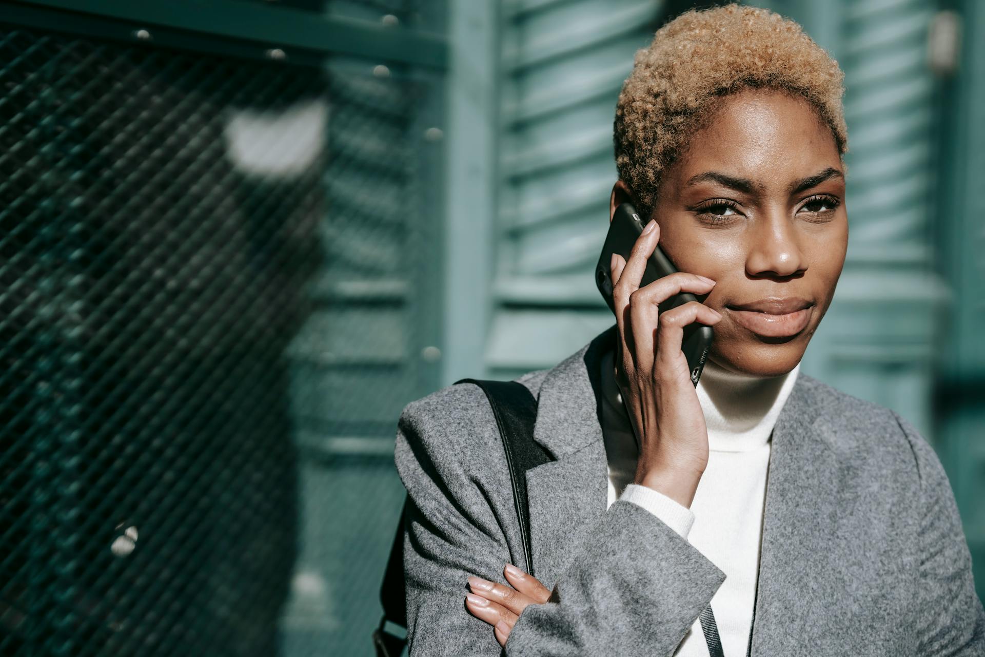 Confident young ethnic female entrepreneur with short Afro hair smiling while having phone conversation on city street on sunny day