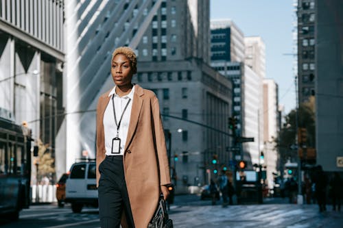 Free Self assured young African American female executive in stylish elegant coat and white shirt with name tag on neck walking on city street with hand in pocket Stock Photo