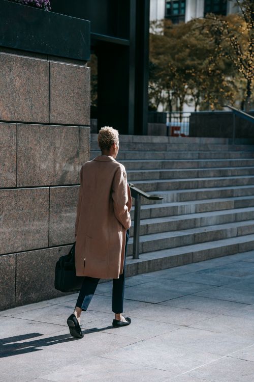 Free Back view full body of unrecognizable female entrepreneur in trendy brown coat with handbag and hand in pocket walking along pavement near office building Stock Photo