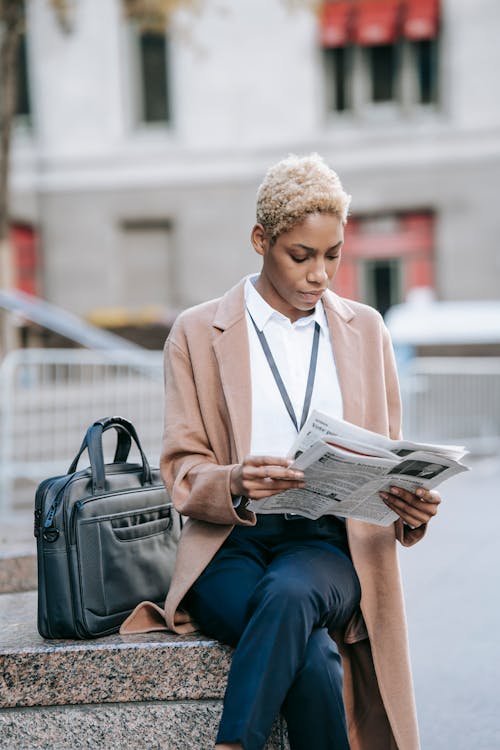 Free Focused young African American female entrepreneur in elegant brown coat sitting on stone border near laptop bag with crossed legs and reading newspaper Stock Photo
