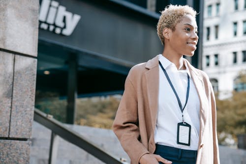 Low angle of smiling African American female entrepreneur in trendy brown coat with badge standing near modern office building and looking away