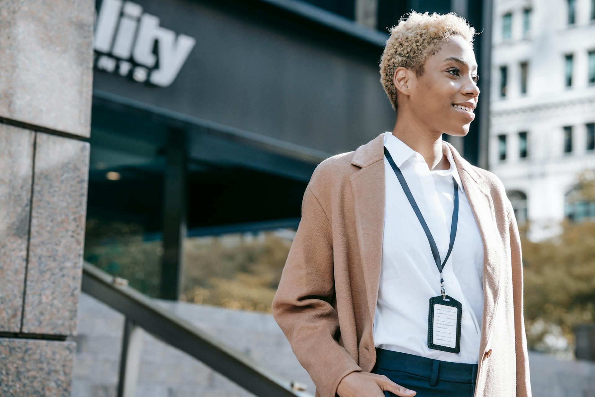 Low angle of smiling African American female entrepreneur in trendy brown coat with badge standing near modern office building and looking away