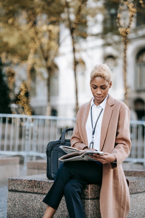 Free Serious young African American female entrepreneur sitting on stone border near laptop bag with legs crossed and reading newspaper with interest Stock Photo