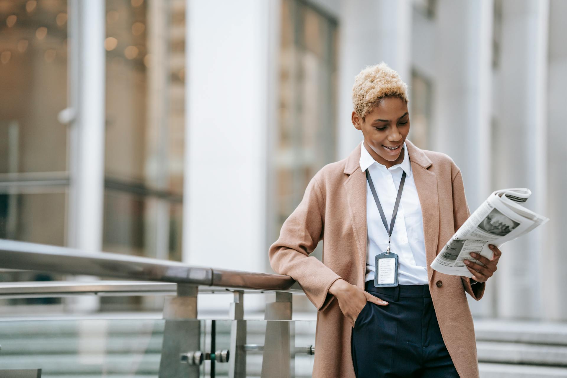 Happy young African American female entrepreneur with badge and hand in pocket in stylish brown coat reading newspaper while standing near stairs leaning on railing