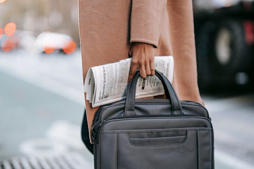 Crop unrecognizable female entrepreneur in stylish brown coat with laptop in bag and newspaper crossing road