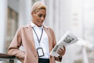 Serious young African American female entrepreneur with badge in elegant brown coat standing near railing and reading newspaper