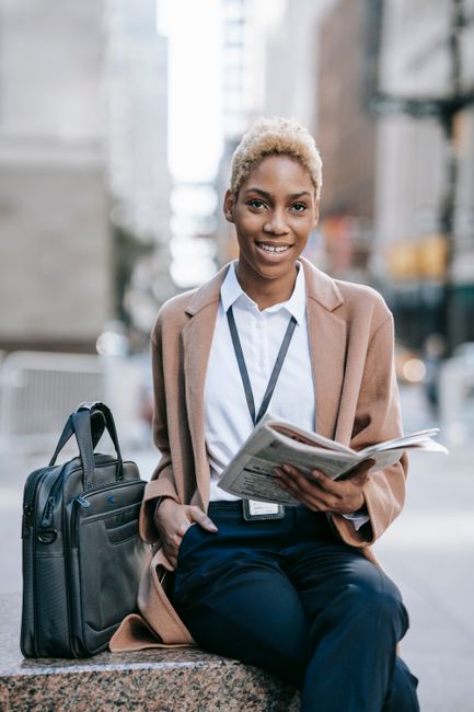 Happy young African American female entrepreneur in classy brown coat sitting on stone bench with crossed legs near bag and looking away while reading newspaper