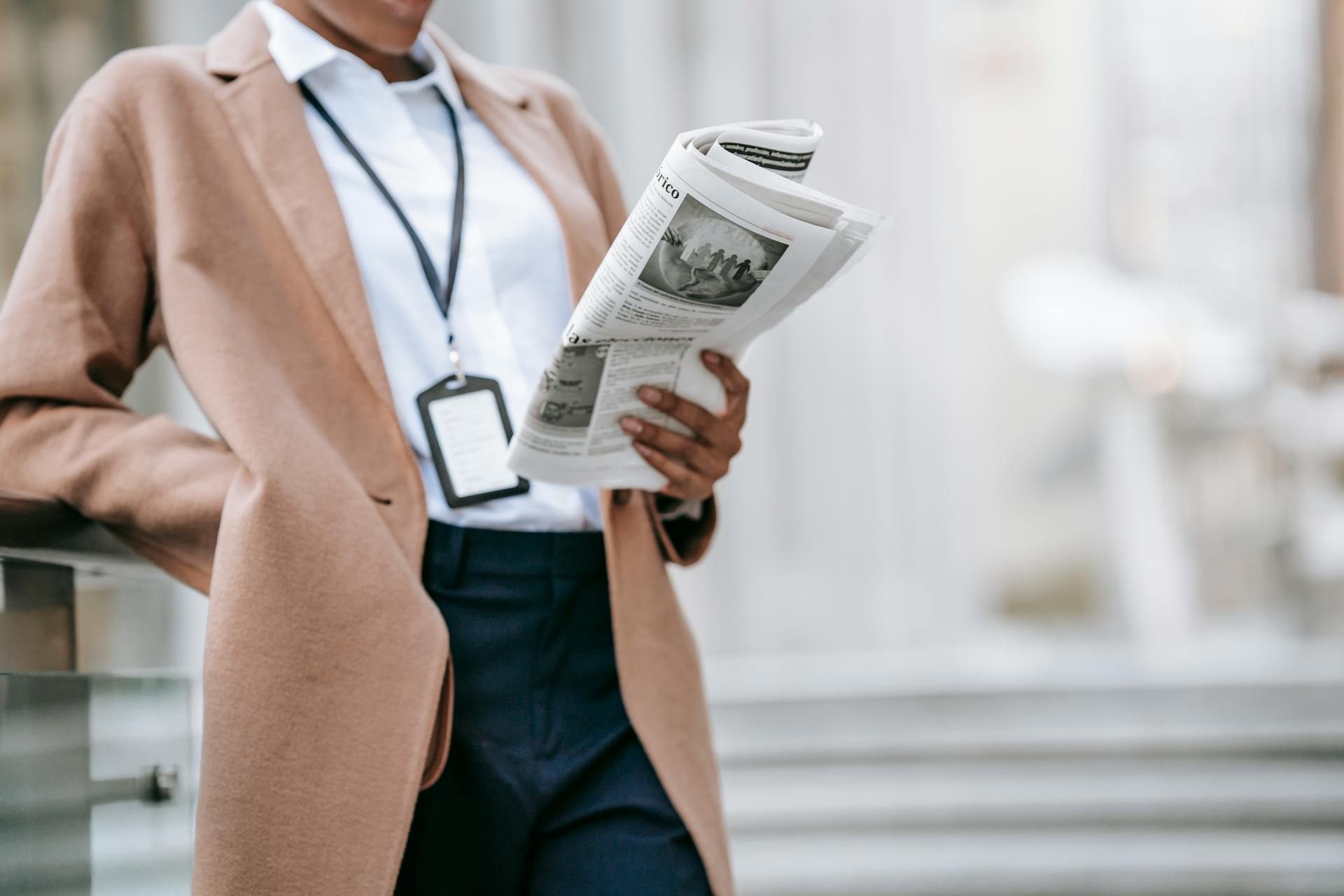 Crop unrecognizable young African American female entrepreneur in elegant brown coat with badge reading newspaper while leaning on railing near stairs