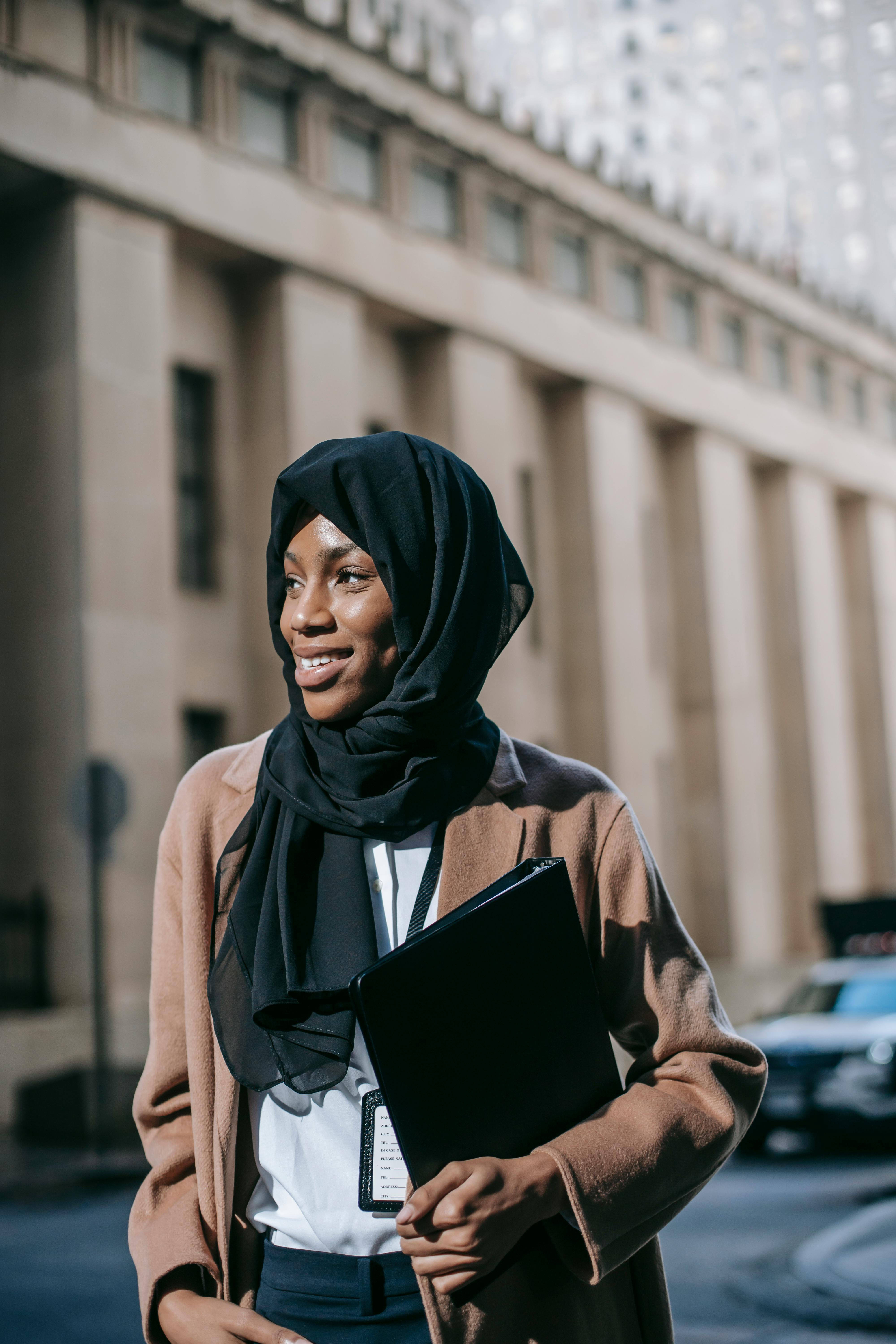 Positive young African American female student in headscarf with folder of documents standing on street against blurred building