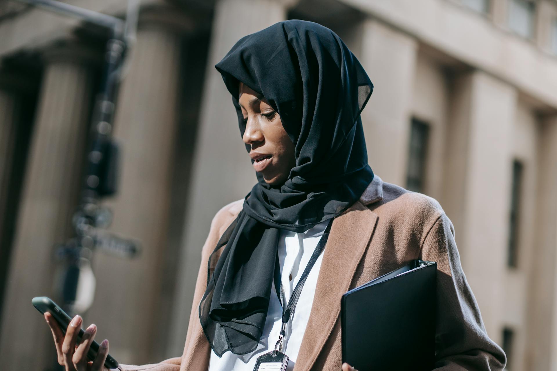 Low angle of focused young African American female in black hijab with folder with project documents chatting on smartphone