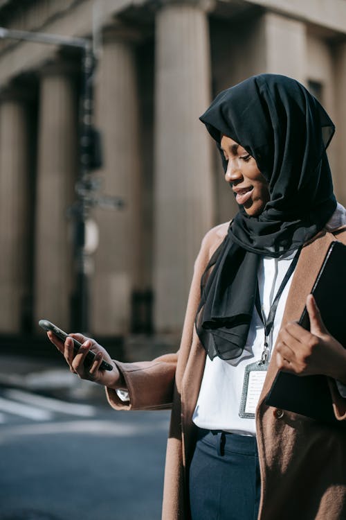 Content young African American female in black headscarf with folder of documents text messaging on phone standing near road
