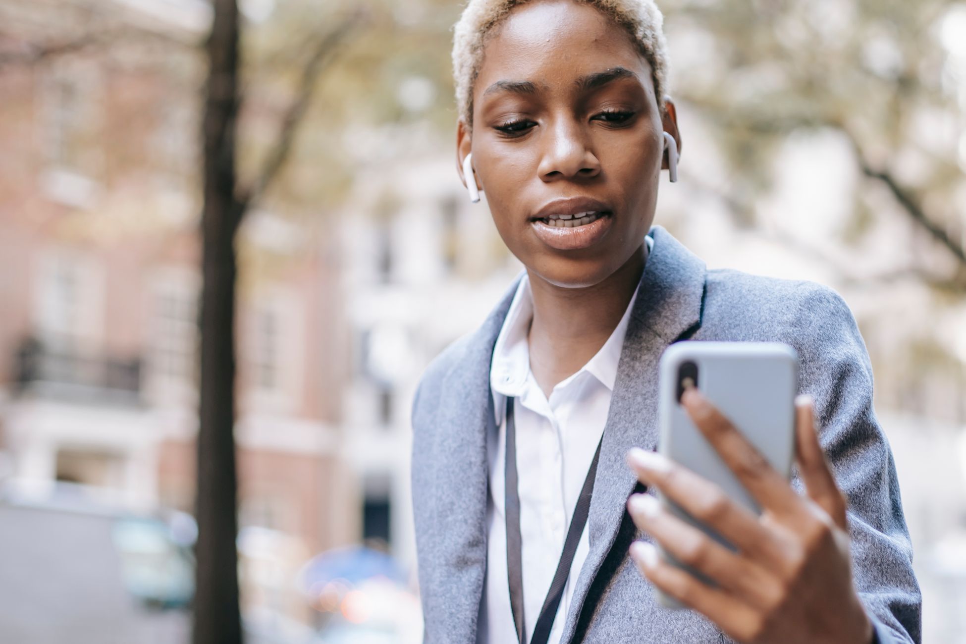 Crop serious African American lady wearing formal clothes and earphones using smartphone for video call in city street