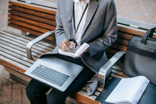 Free From above of crop faceless entrepreneur wearing formal clothes taking notes while sitting with laptop on wooden bench in daytime Stock Photo