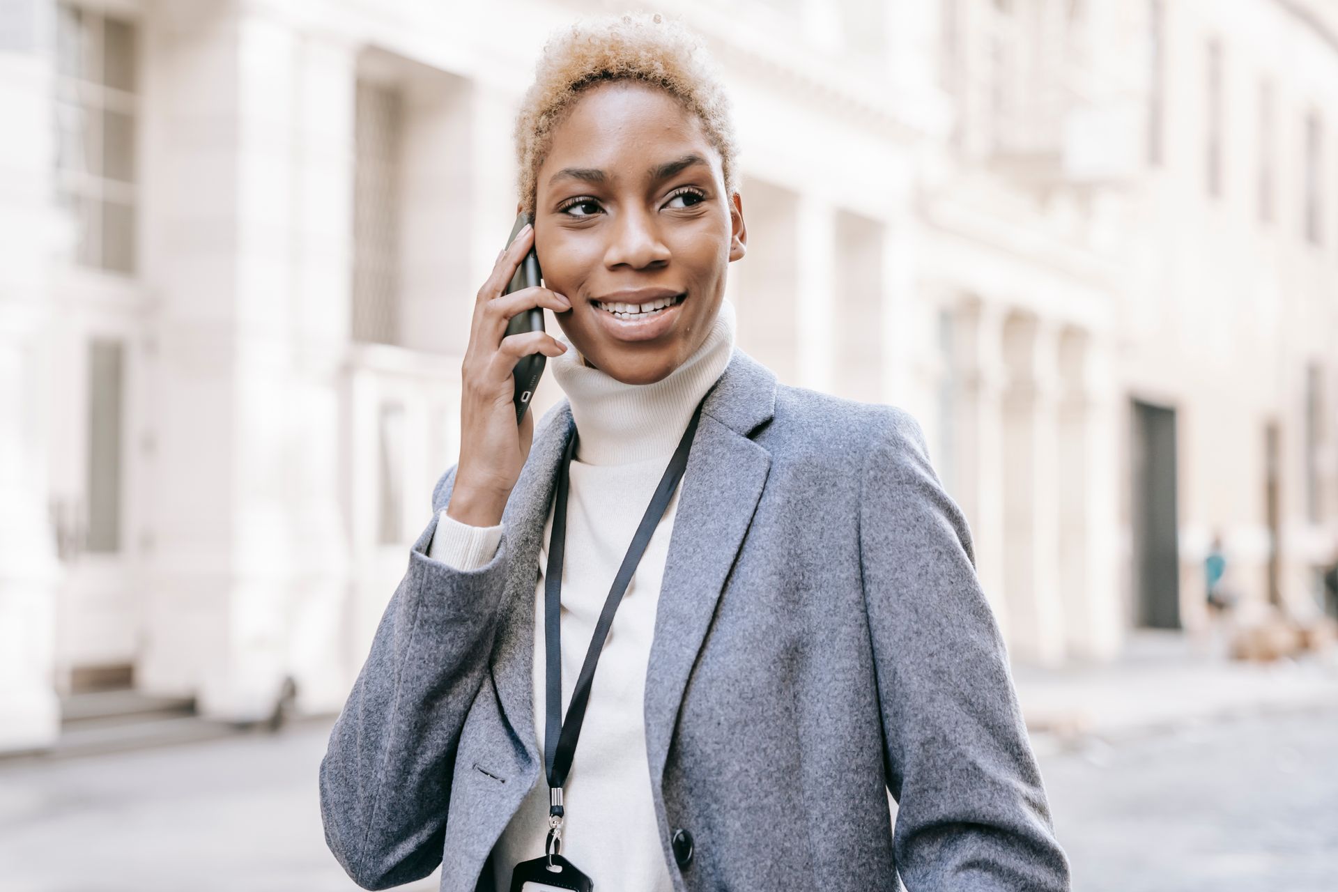 Delighted African American lady wearing white turtleneck talking on mobile phone while looking away on urban background