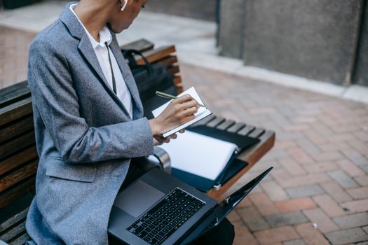 From above crop confident African American businesswoman wearing classy coat rewriting information from documents while working remotely with laptop in daylight