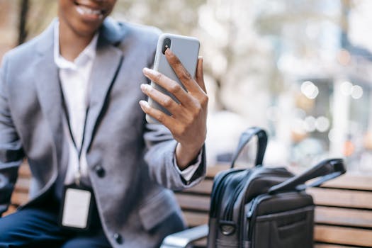 Crop unrecognizable happy young African American female employee in formal outfit and name tag smiling while having video call on mobile phone sitting on bench on city street