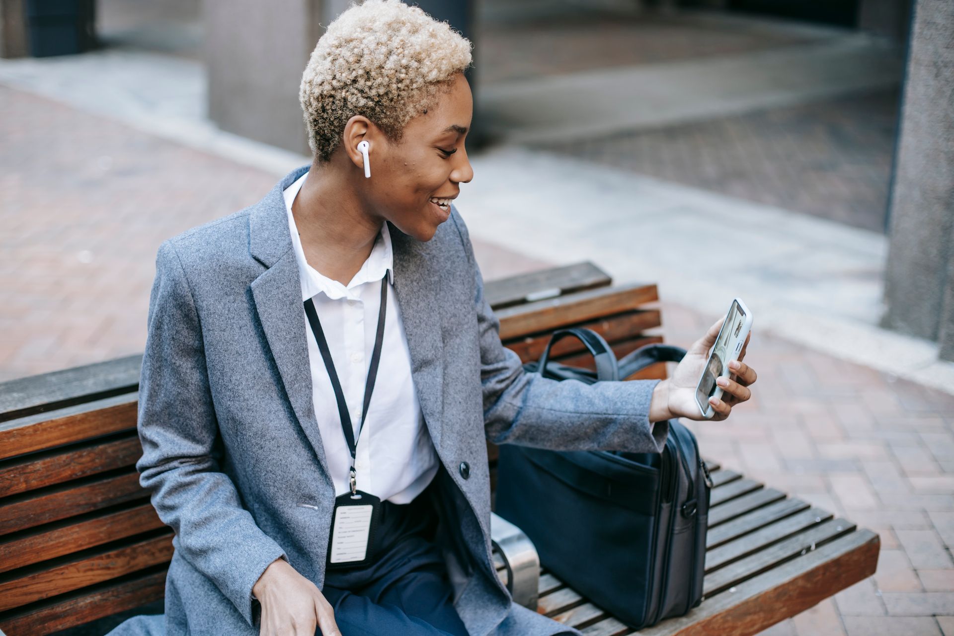 From above of positive young stylish black female entrepreneur in formal clothes and TWS earphones sitting on wooden bench on street and smiling while having video call on smartphone