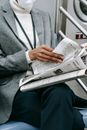 Crop anonymous black female entrepreneur in elegant outfit and medical mask reading newspaper while riding train