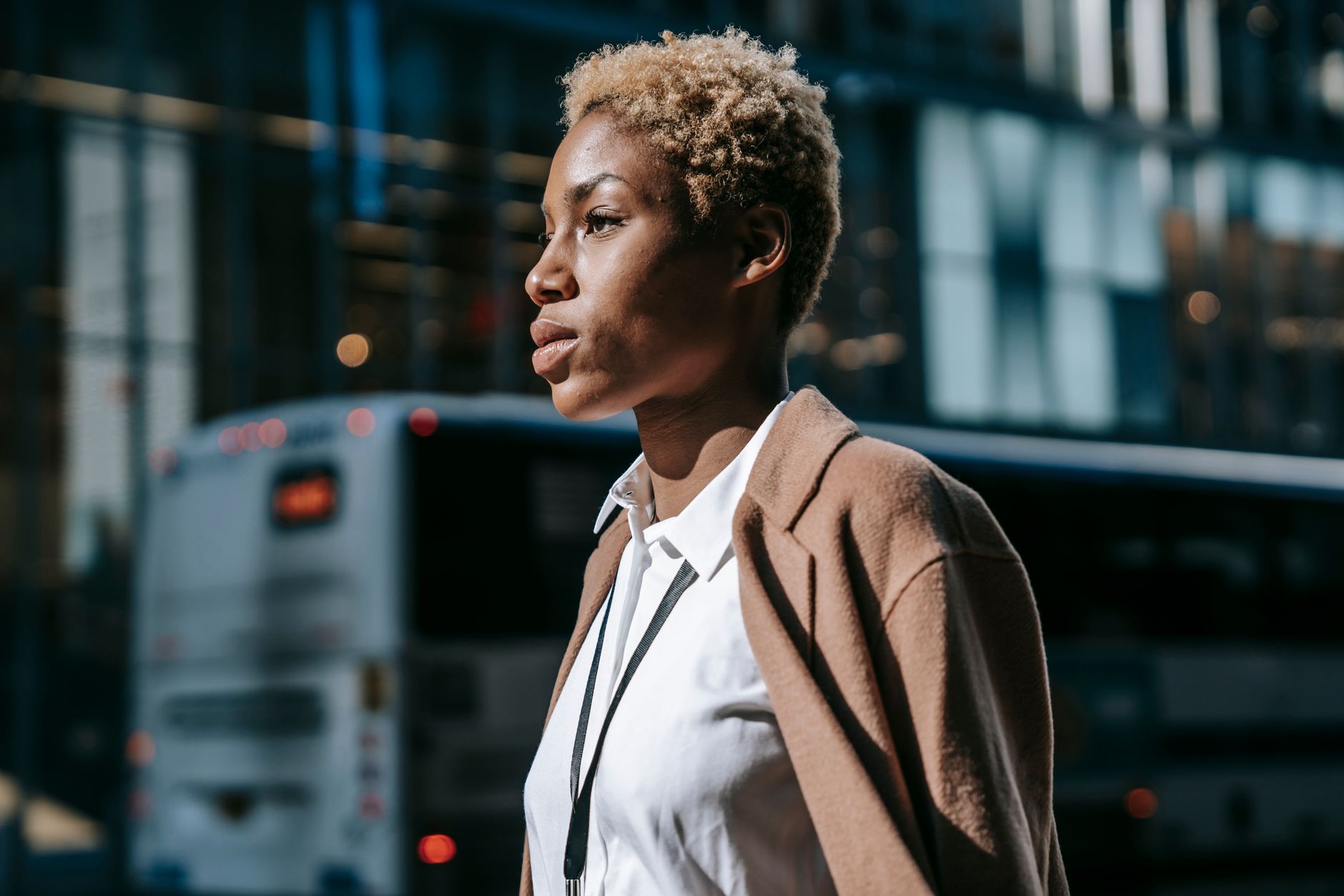 Side view of serious young African American businesswoman with short blond hair in white shirt and coat walking on city street on sunny day