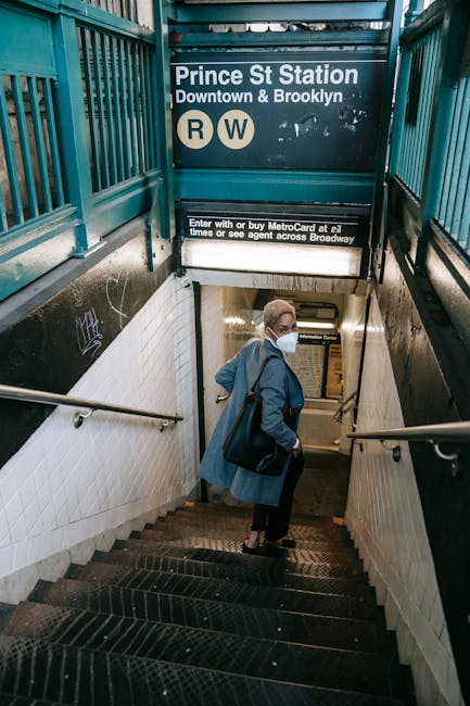 From above side view of anonymous young ethnic female with short blond hair in classy outfit and medical mask walking downstairs towards subway station entrance and looking over shoulder