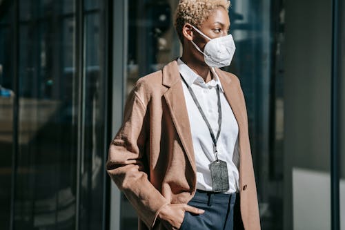 Crop anonymous self assured African American businesswoman in elegant outfit and protective mask standing on street with hand in pocket and looking away during coronavirus pandemic