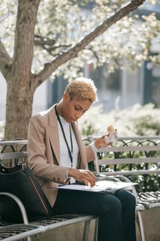 Concentrated young African American female employee with short blond hair in formal outfit sitting on bench in city park and reading documents during lunch break