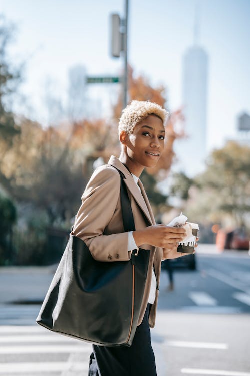 Free Side view of positive young African American female entrepreneur with short blond hair in elegant outfit drinking takeaway coffee and eating sandwich while walking on city street during break Stock Photo