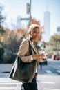 Side view of positive young African American female entrepreneur with short blond hair in elegant outfit drinking takeaway coffee and eating sandwich while walking on city street during break