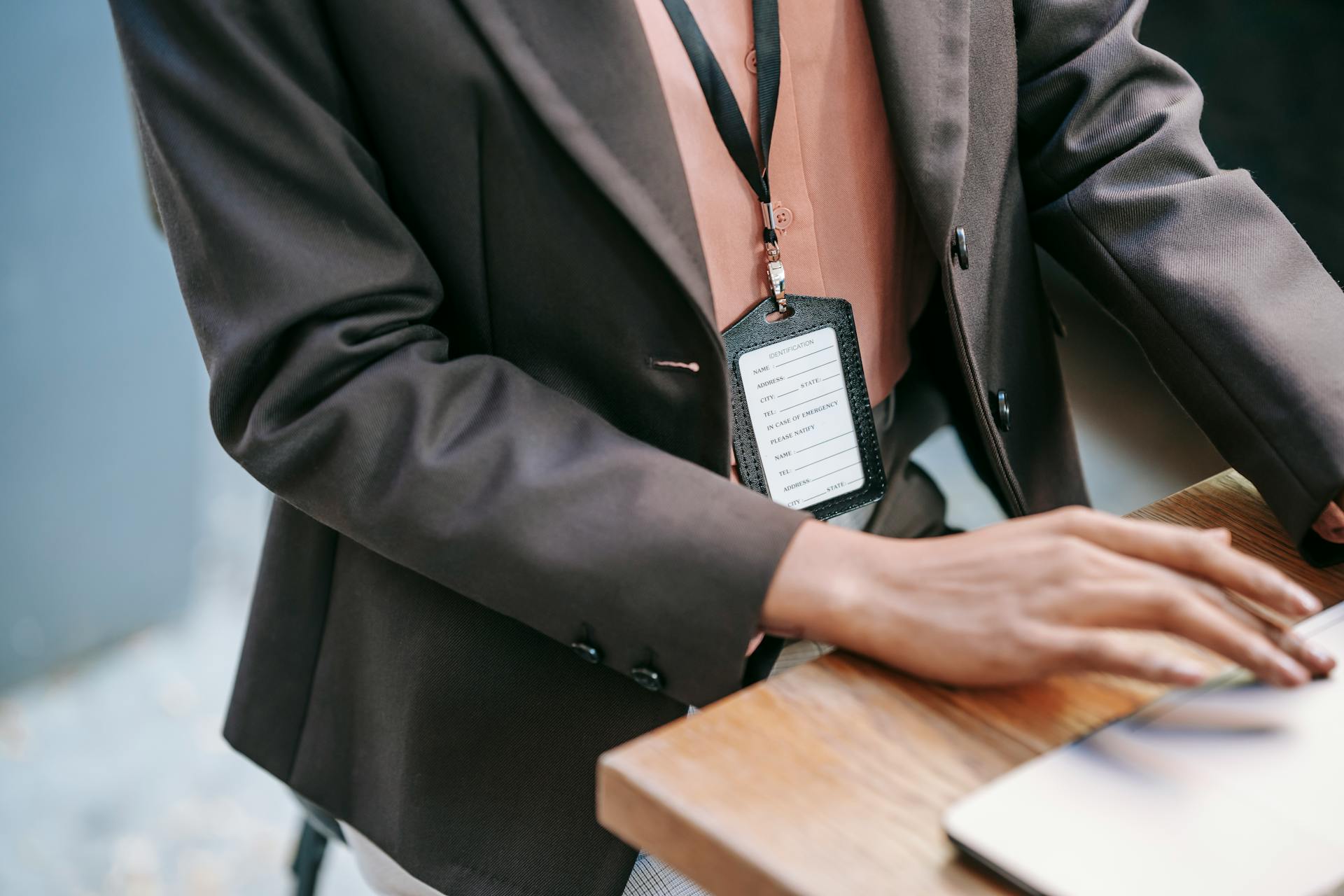 From above of crop anonymous female entrepreneur in formal clothes and name tag on neck sitting at wooden table and typing on laptop during work in office