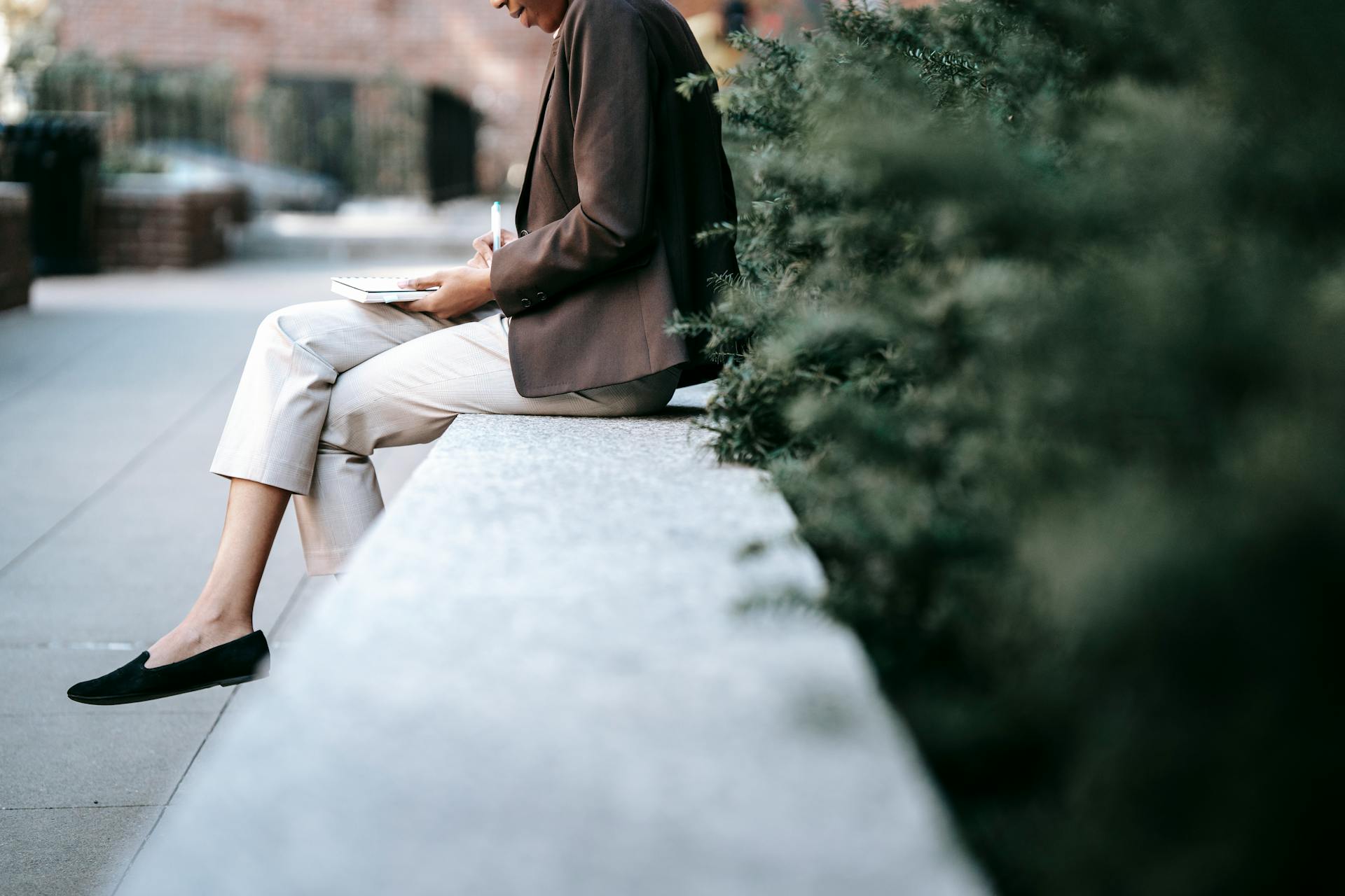 Side view of crop unrecognizable young ethnic female employee in classy outfit sitting on stone border in city park and taking notes in planner