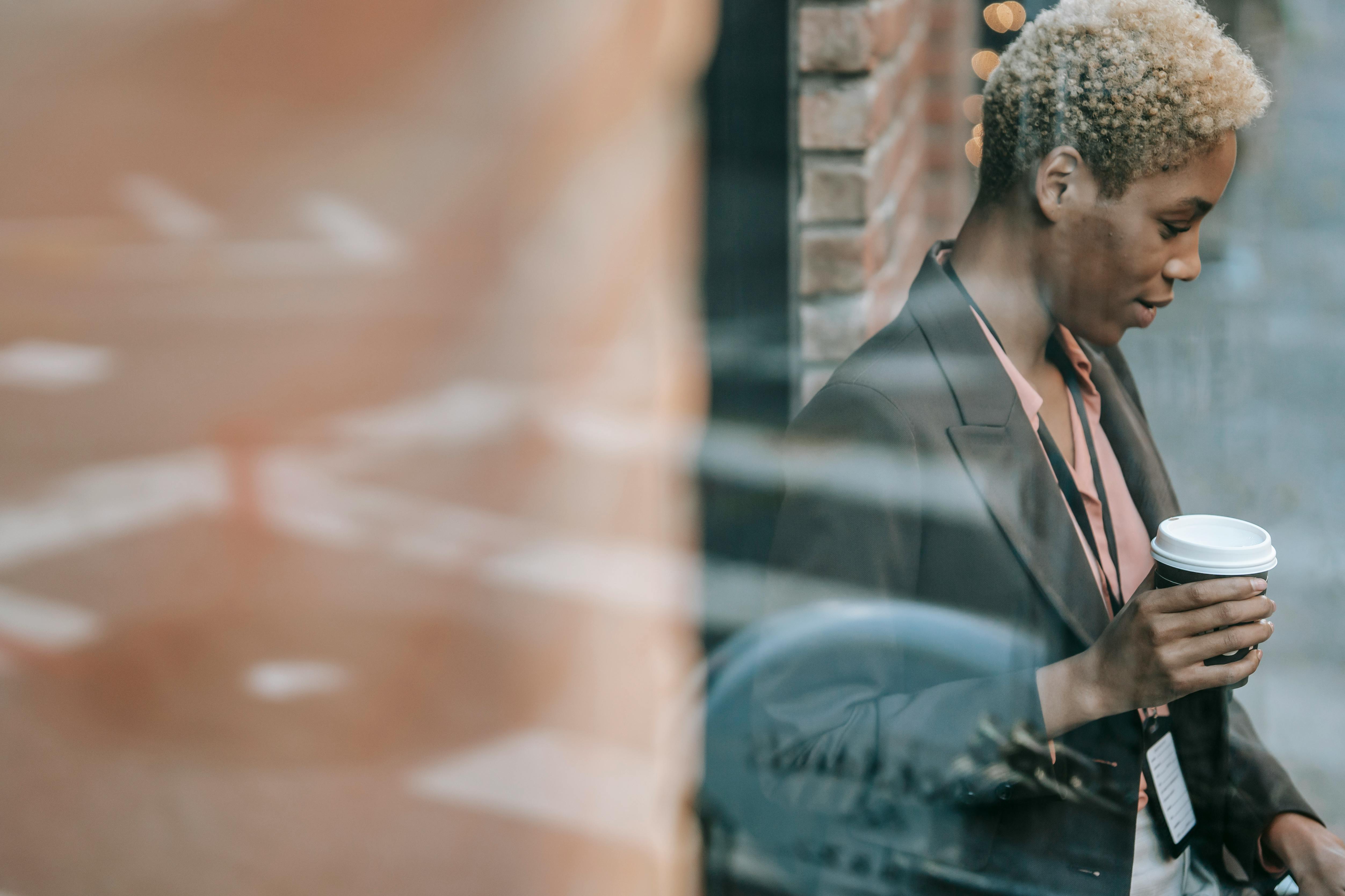 Through glass of serious young African American female manager with short blond hair in classy outfit drinking takeaway coffee in street cafe