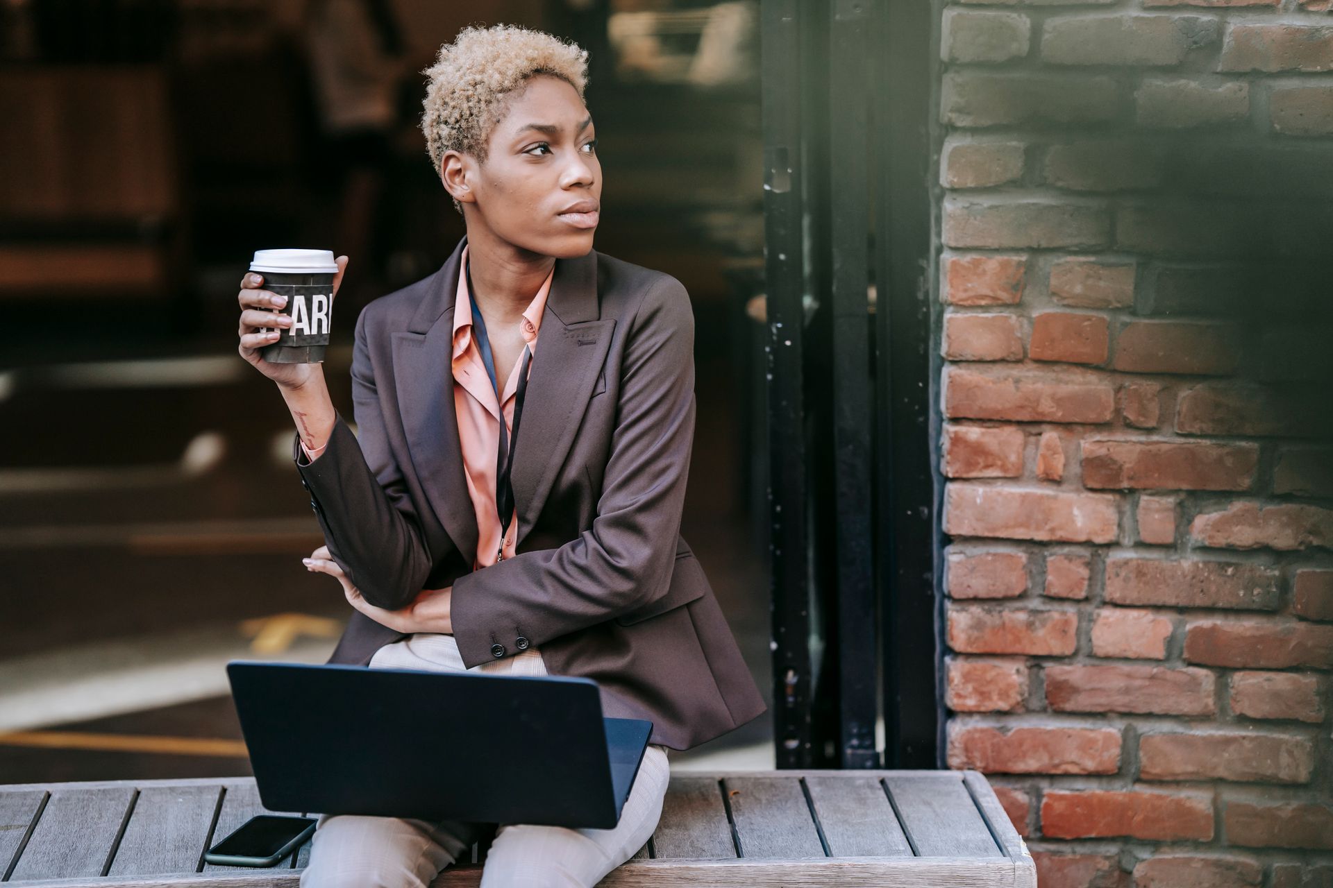 Dreamy young ethnic female manager in classy outfit drinking takeaway coffee and looking away thoughtfully while sitting on bench on city street with laptop during break
