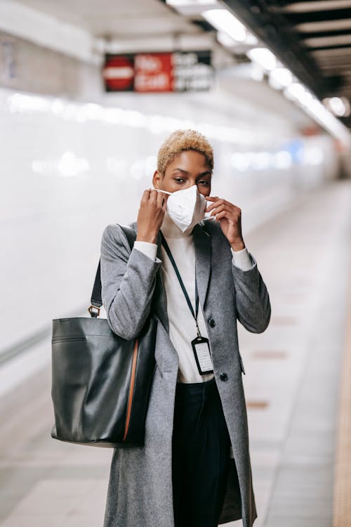 Young African American female in stylish formal wear putting on protective mask while waiting for train on underground platform