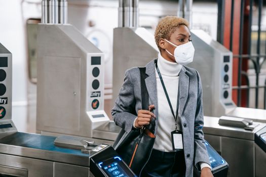 Calm attractive African American female in stylish formal wear and protective mask passing through turnstile gates in underground station