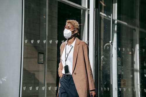 Free Contemplative young African American businesswoman in formal clothes and face mask walking along modern building glass wall on sunny autumn day Stock Photo