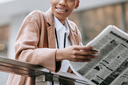 Free Crop happy African American businesswoman in formal clothes reading newspaper and looking away with smile while leaning on railing outside blurred modern building Stock Photo