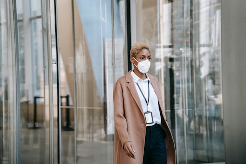 Serious black businesswoman in mask walking along glass building